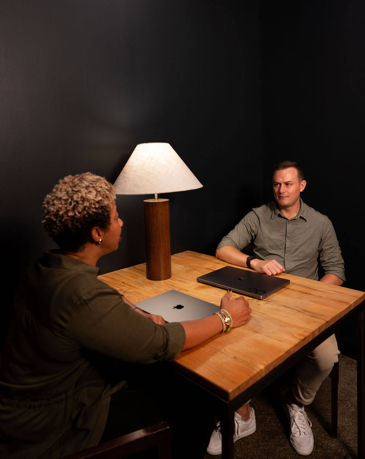Two employees seated at a table with closed laptops in front of them. They are conducting a 1-on-1 meeting.