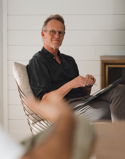 A man sits in a chair with his legs crossed smiling