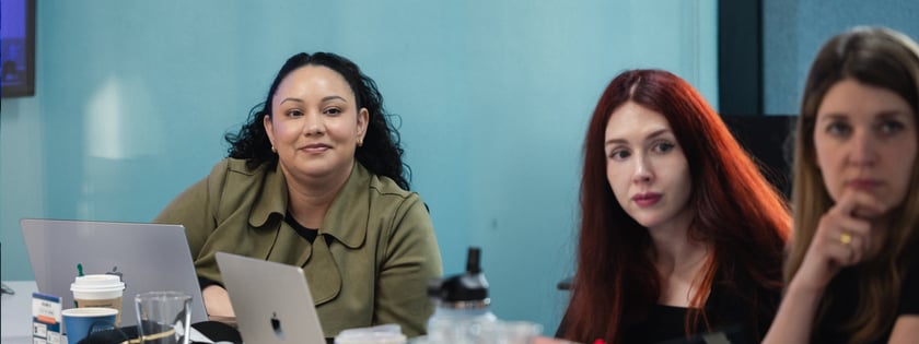 A group of three women sit at a table in front of Macbooks while looking toward the camera