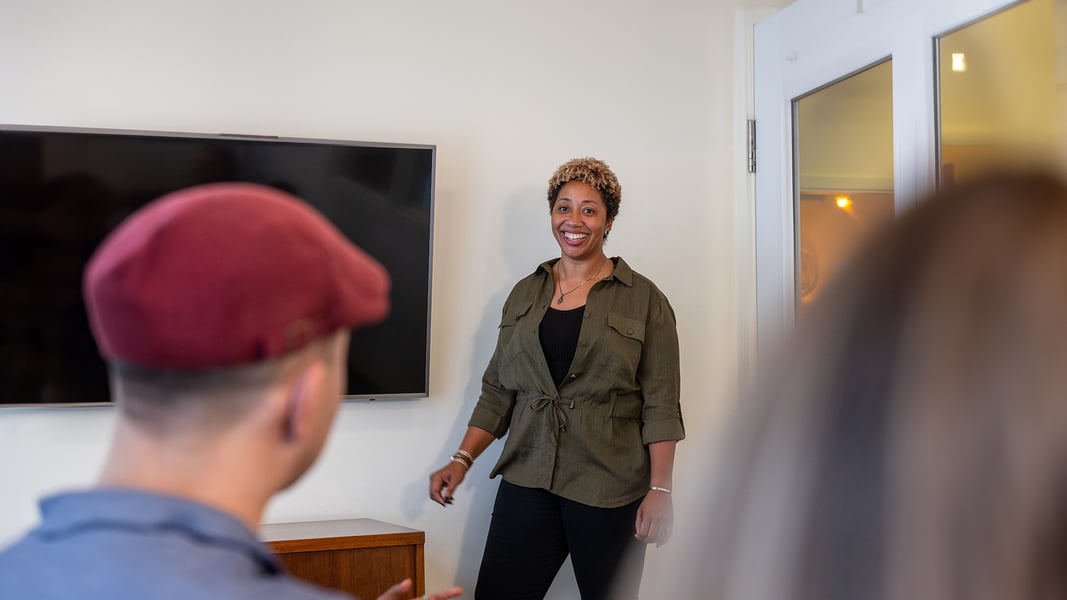 A woman stands in front of a room smiling while several people look on A woman stands in front of a room smiling while several people look on