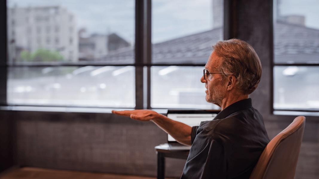 A man sits in a chair in front of a window. He holds his hand, palm down, out in front of him.