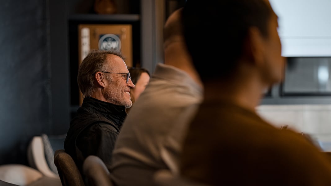 A man smiles as he sits at a table with others to the left and right of him. 