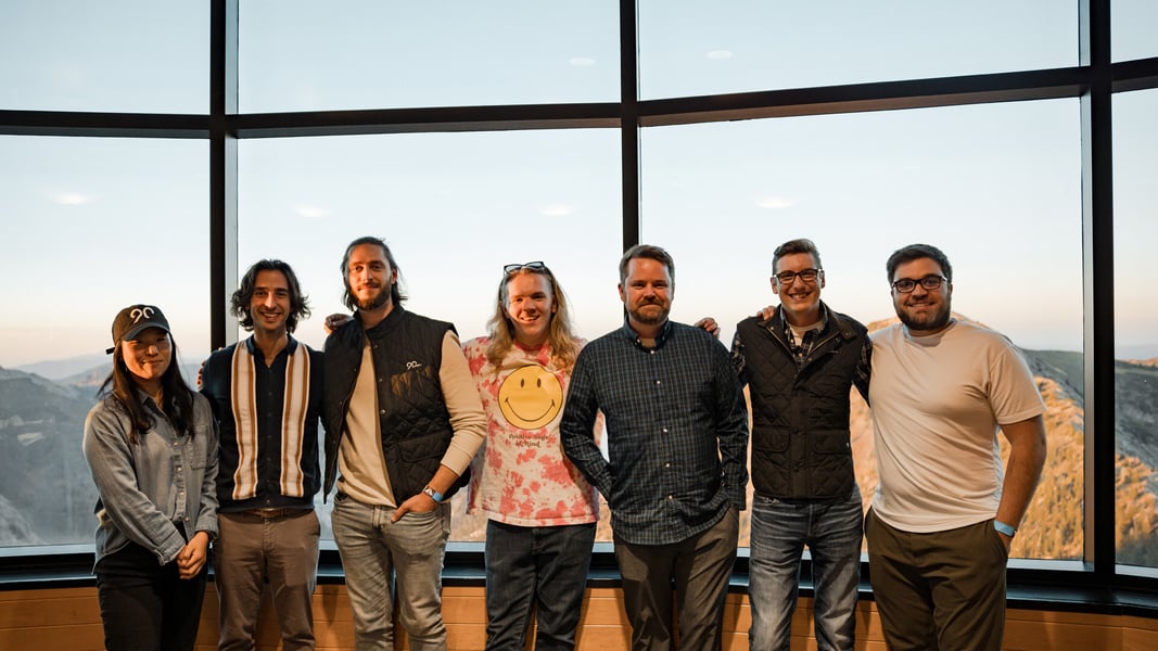 A group of seven people stand in front of a picture window with their arms around each other smiling at the camera