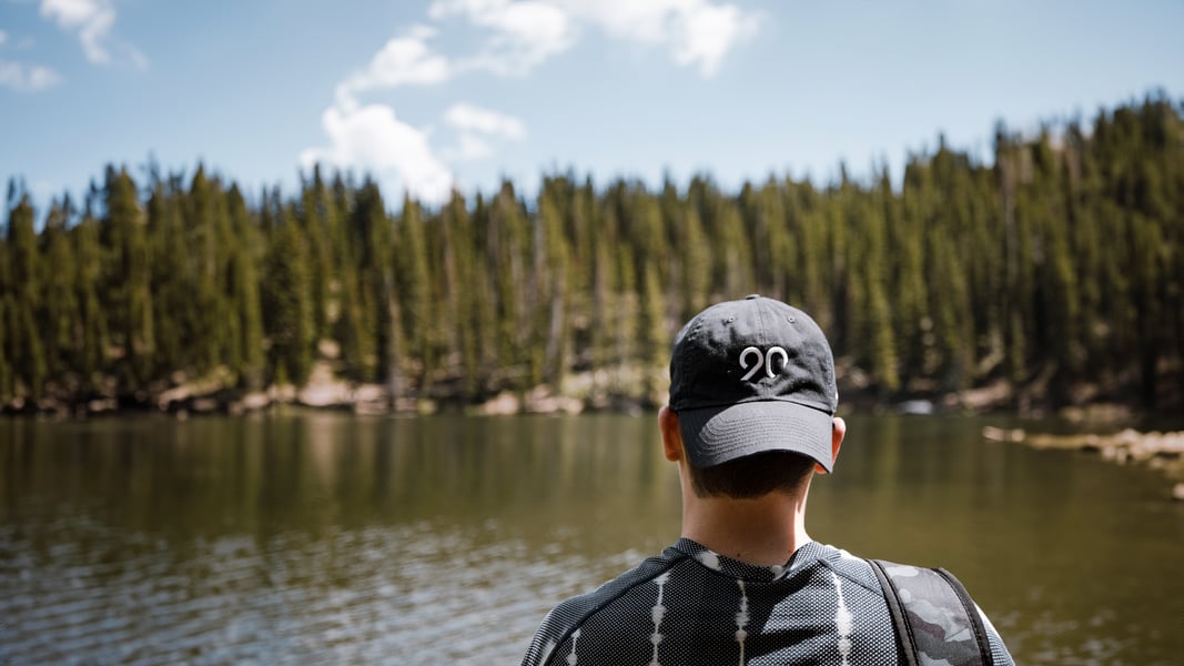 A man wearing a backwards black baseball cap with the Ninety logo on it looks toward a river. There is a line of evergreen trees on the opposite bank