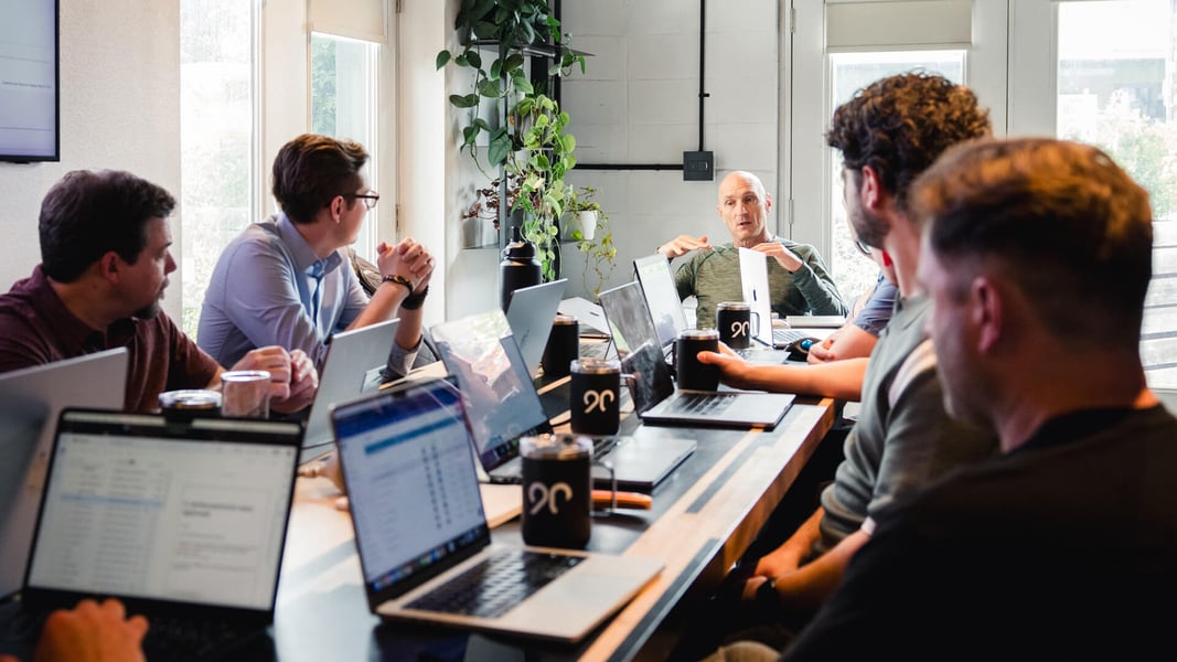 Several men sit around a long rectangular table with laptops in front of them. Several men sit around a long rectangular table with laptops in front of them.