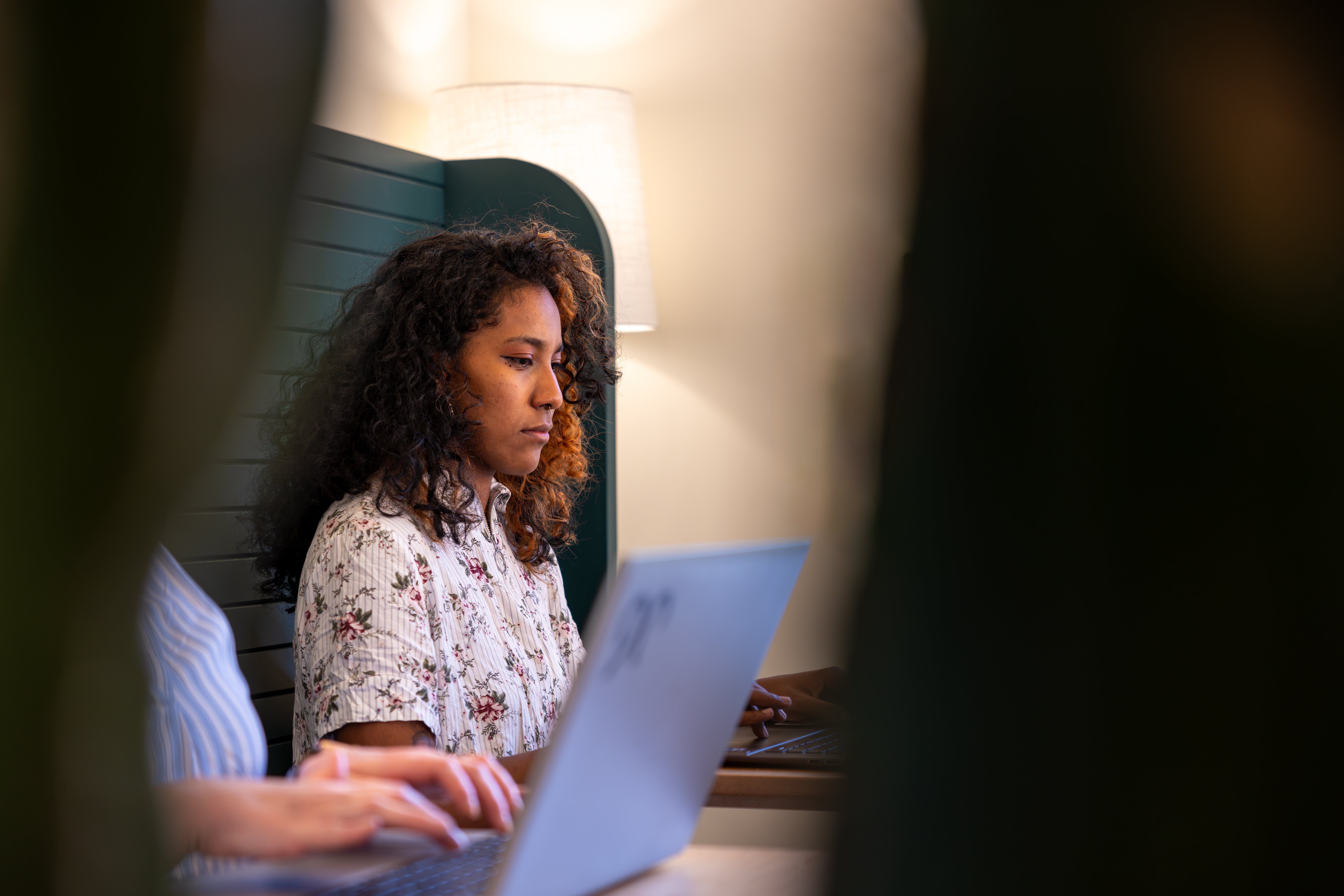 A woman sits and looks at her computer. Another person is on a computer next to her. 