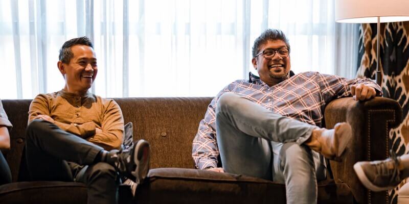 Two men sitting on a brown couch in a room with curtains and a lamp, both smiling and casually dressed.