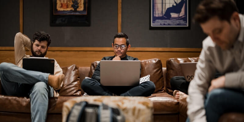 Three people sitting on a brown leather sofa, each focused on a laptop. Two are on either end, and one is in the center. A blurred person is in the foreground. Wall art is visible behind them.