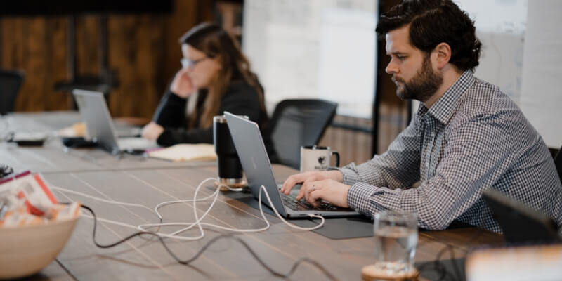 Two people working on laptops at a conference table with various items, including a bowl of snacks, a mug, and a glass of water.