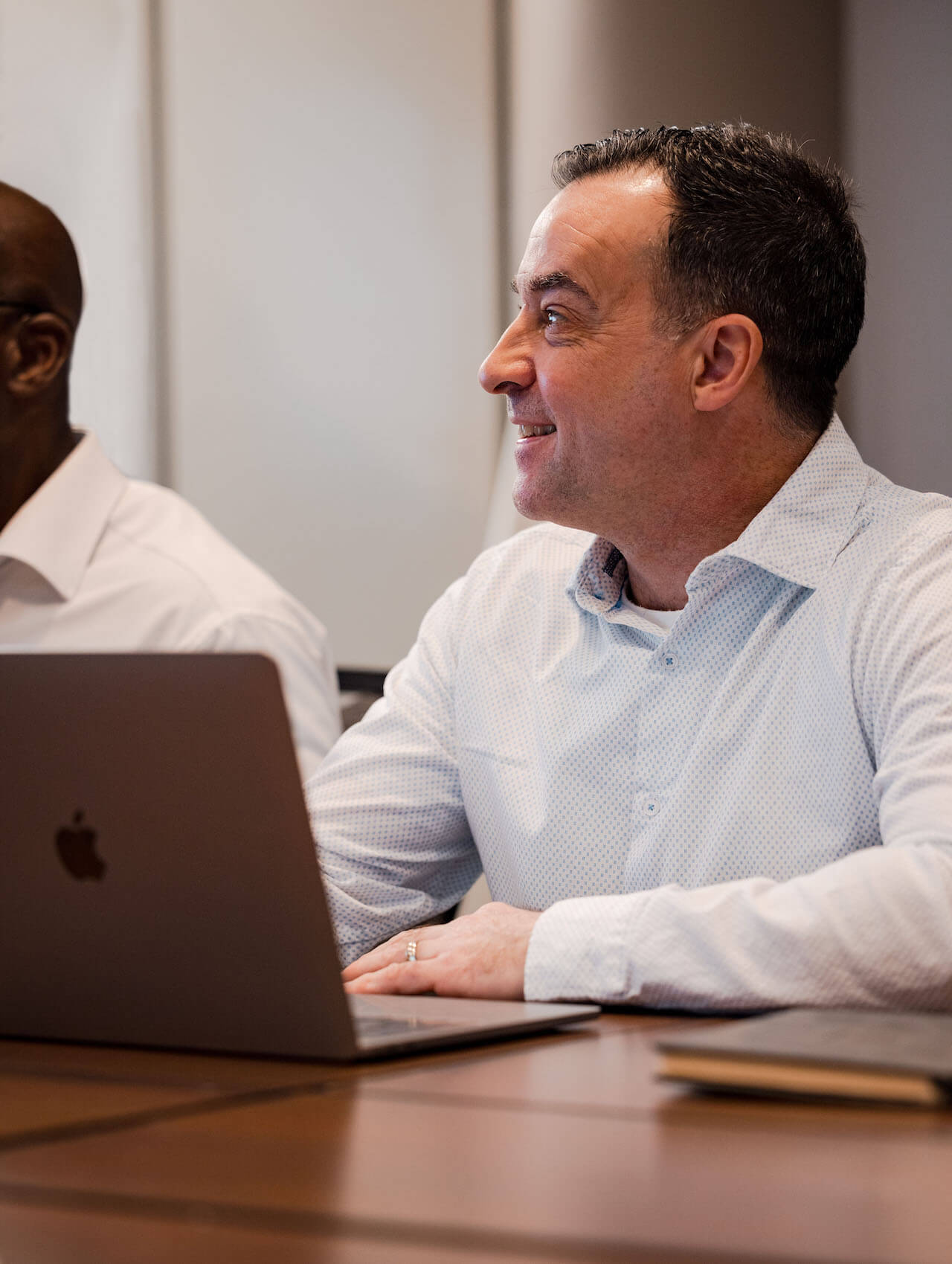 A man wearing a white shirt in a meeting, listening to someone speaking.