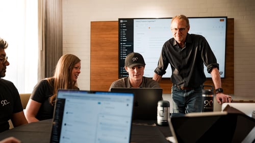 A man stands with a large screen behind him as others sit at a conference room table with laptops.