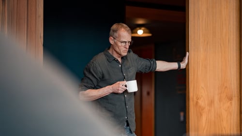 A man stands with a mug and leans on a door frame.