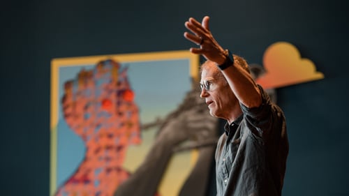 A man stands with a stern expression and his hand raised to the side.