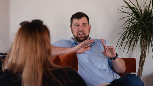 A man sitting down, using hand gestures while talking to a woman at work.