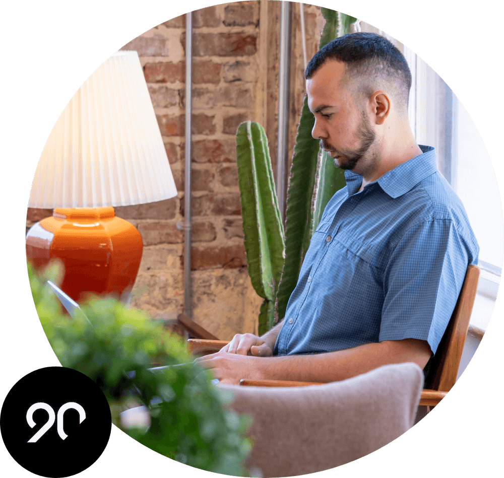 A man seated in a cafe working on his laptop. The Ninety logo is in the corner.