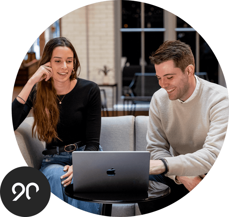 Two employees having a meeting using a shared laptop. Superimposed is the Ninety logo. Two employees having a meeting using a shared laptop. Superimposed is the Ninety logo.