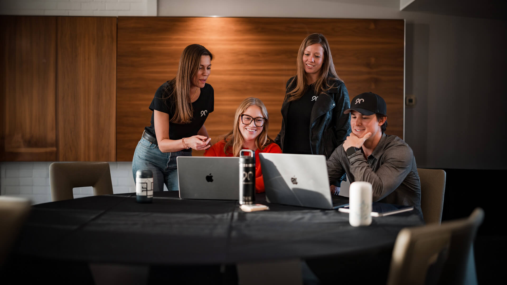 Four people look at 2 computers on a table and smile.