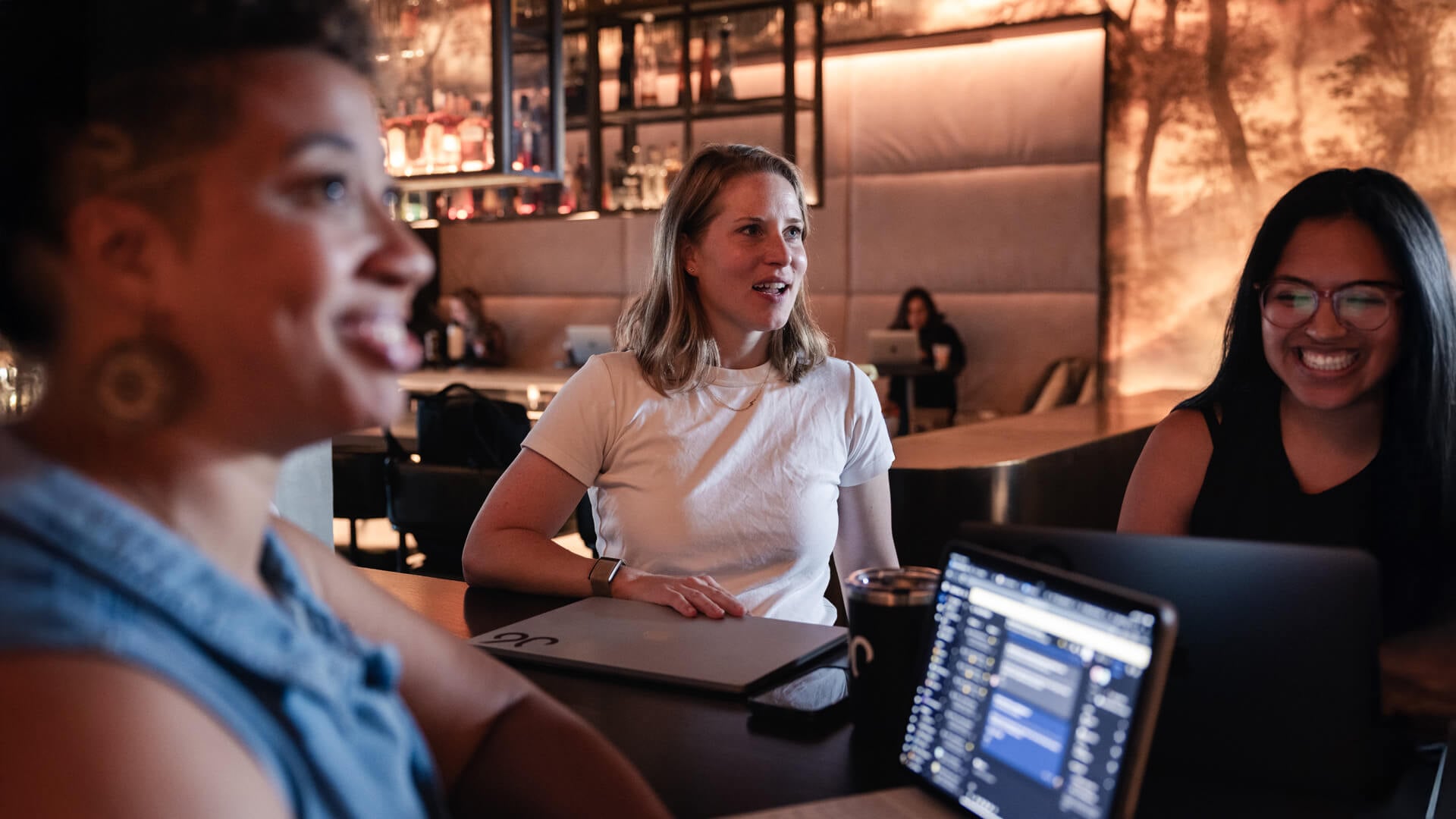 Three women sitting down at a table talking and working on laptops. 
