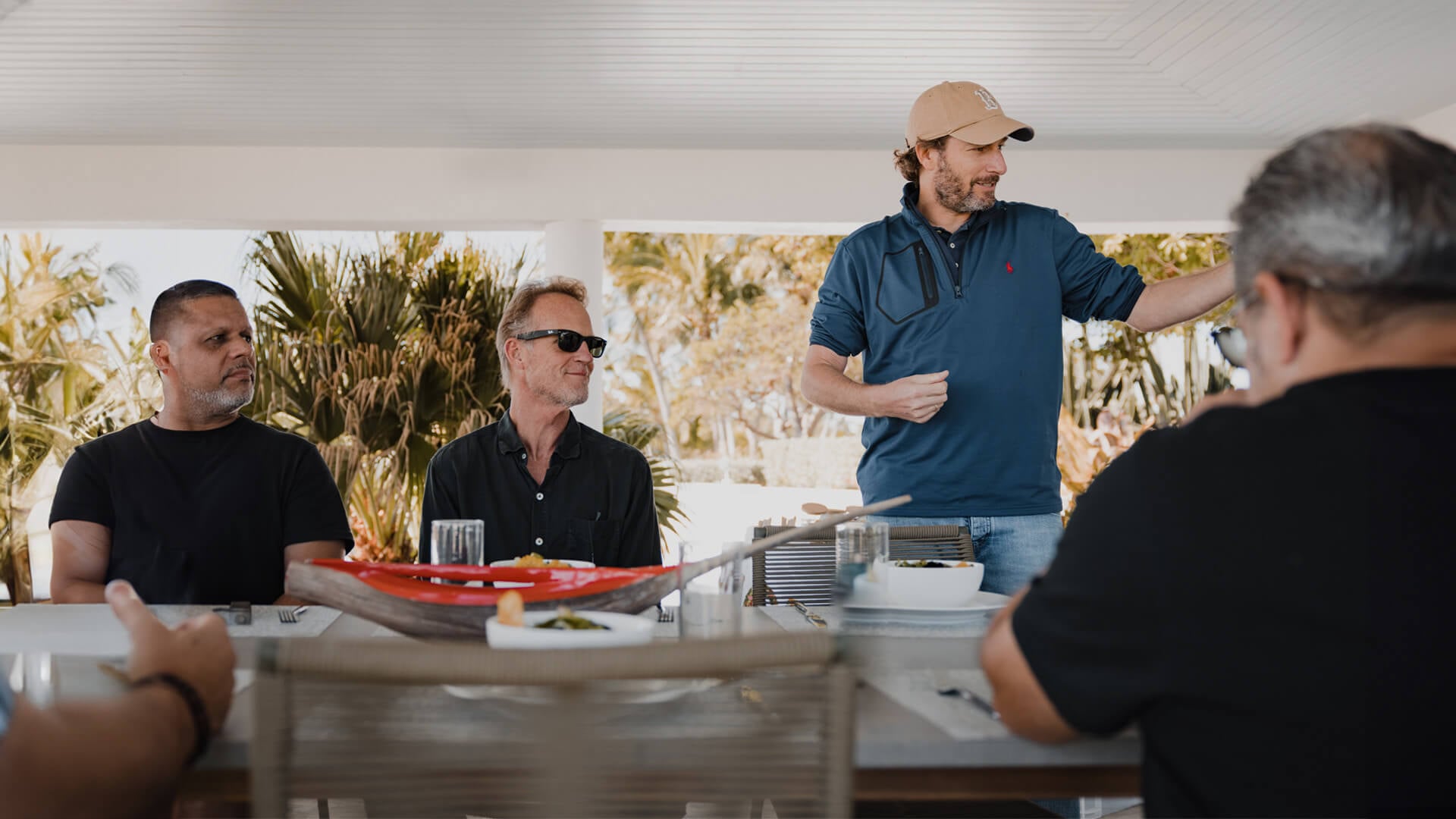 Several men sit outdoors at a table and one stands. 