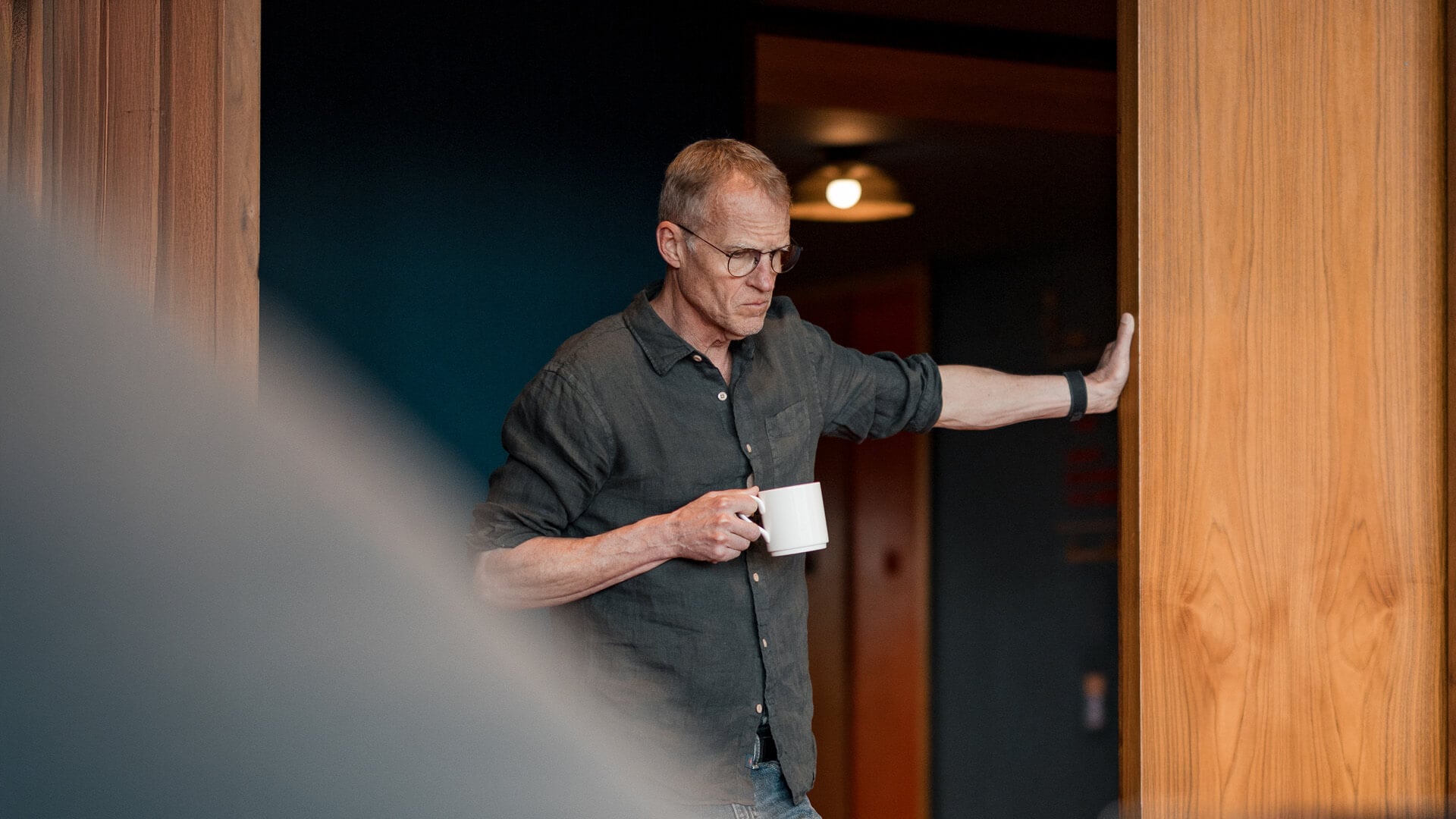 A man stands with a mug and leans on a door frame.