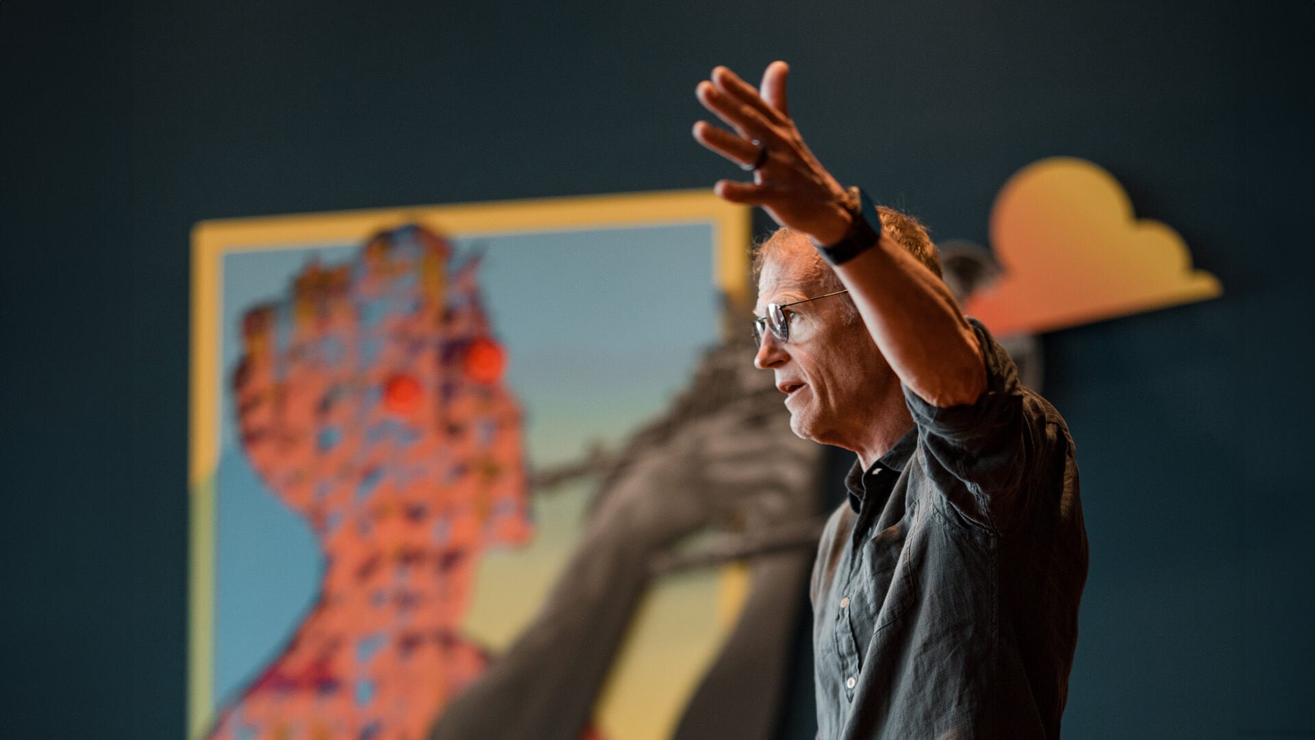 A man stands with a stern expression and his hand raised to the side.