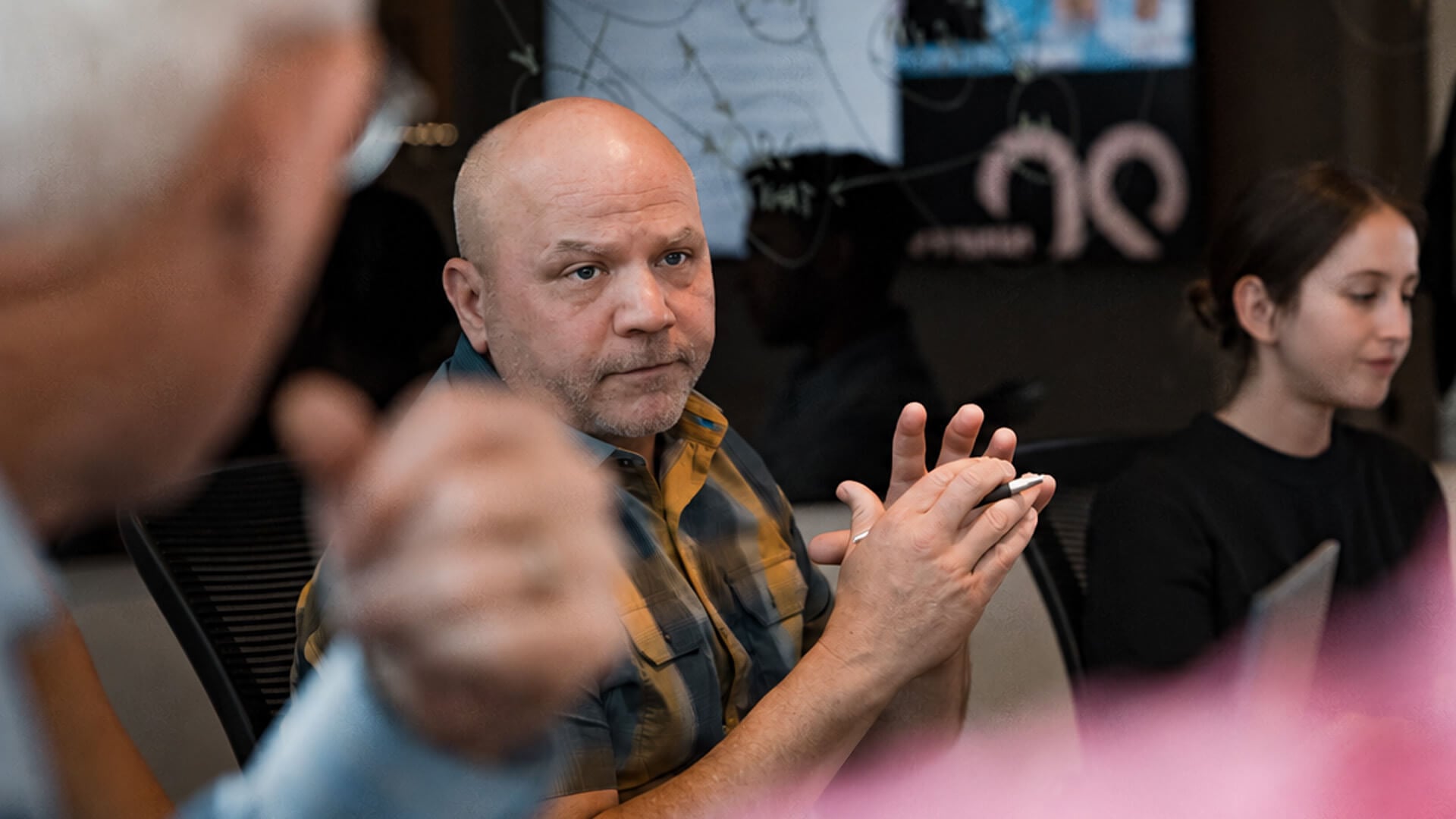 A man with a serious expression has his hands raised in front of his chest. Other people sit around him.