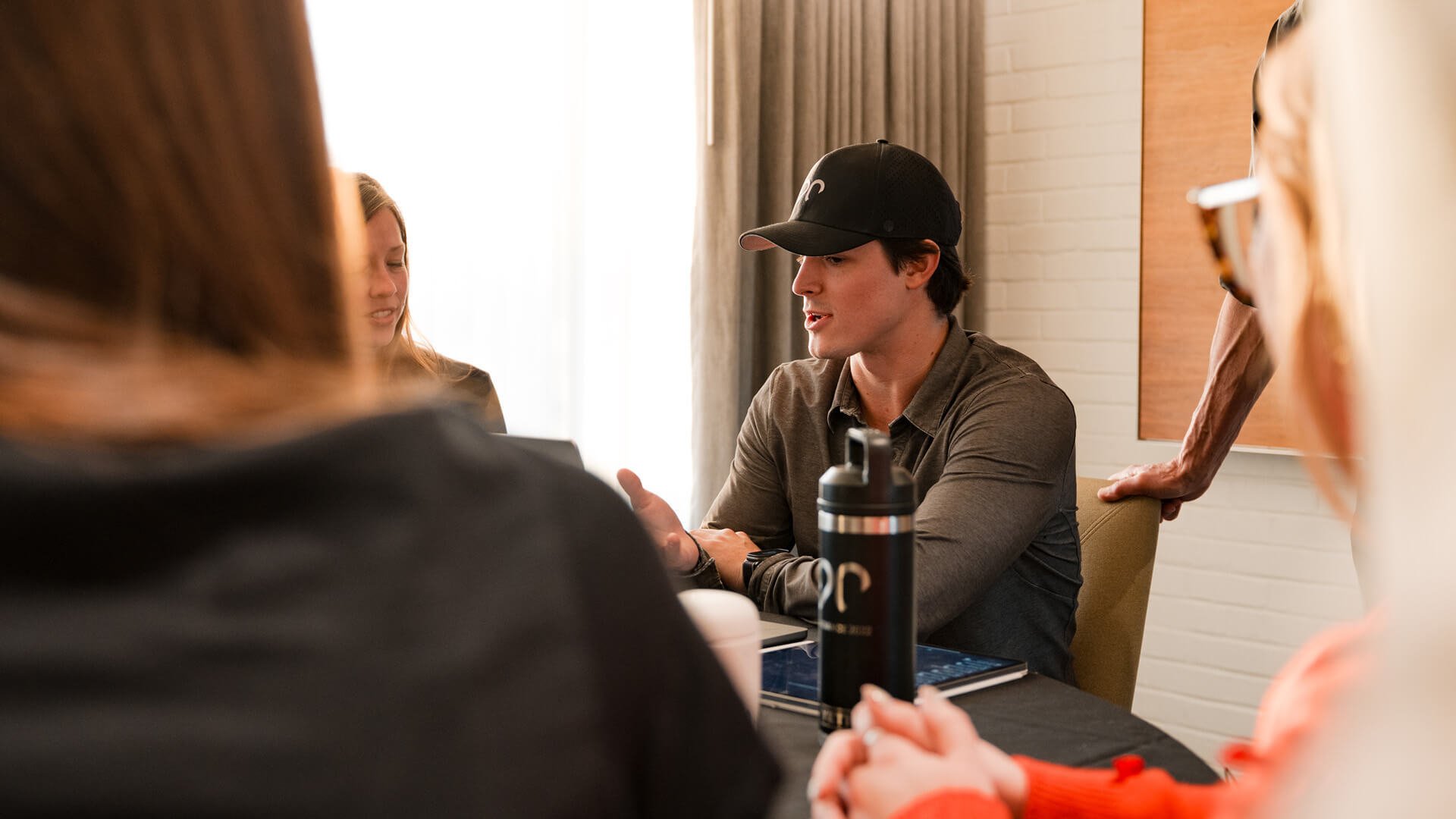 A man wearing a hat sits at a table and talks to other people at the table.