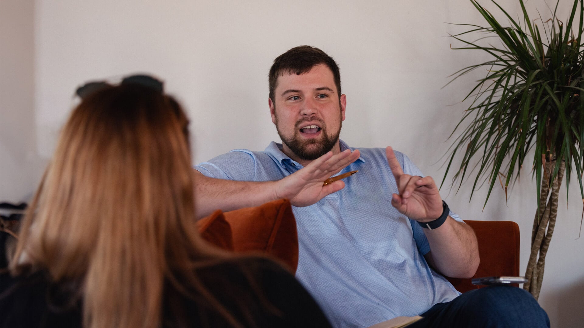A man sitting down, using hand gestures while talking to a woman at work.
