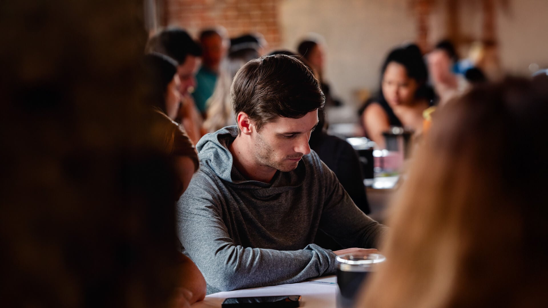 A man sits at a table and looks down. Several other people are seated around him.