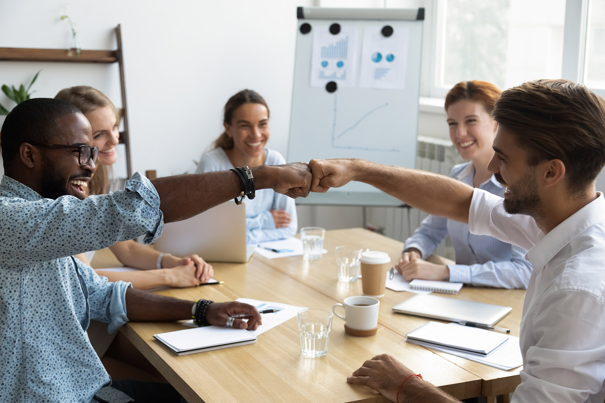 A team of people fist bumping and sitting around a conference table.