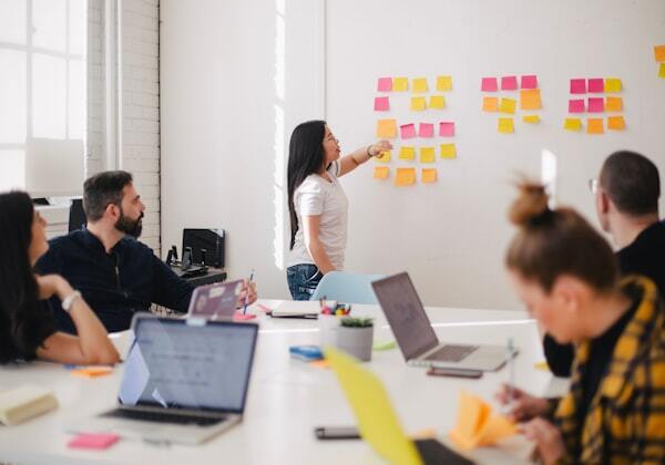 A team collaborates in a conference room, with one person organizing colorful sticky notes on a whiteboard while colleagues review laptops at the table.