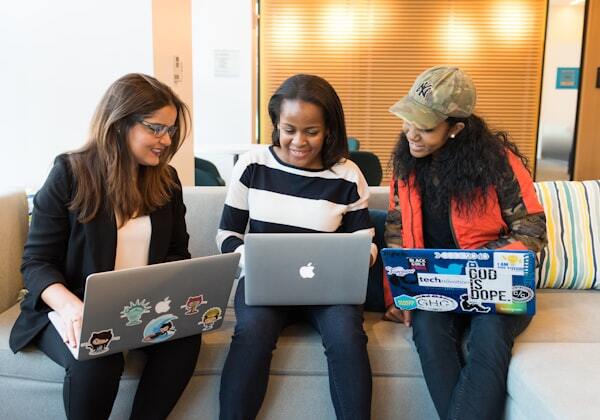 Three women sit together on a couch, smiling and looking at their laptop screens during a casual work session.