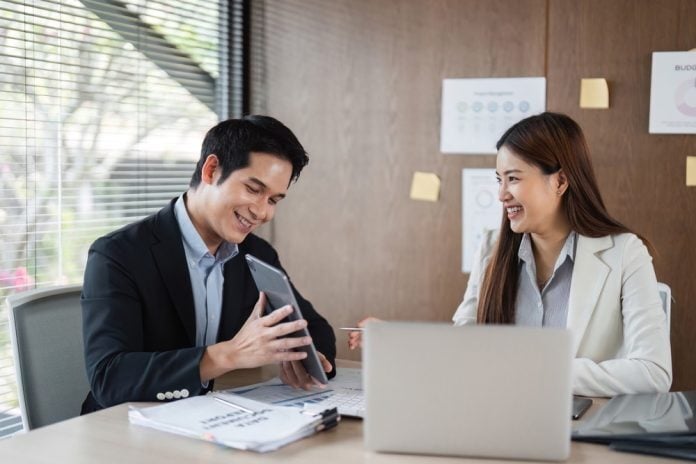 Two smiling business professionals in an office discussing content on a tablet, with a laptop and documents on the desk in front of them.